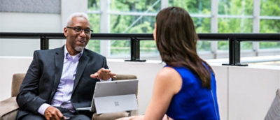 A man in a suit sitting at a table with a laptop and a woman in a blue dress talking to him.