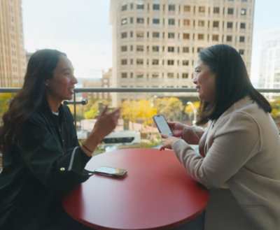 A woman with black hair holding a phone and talking to another woman at a table.