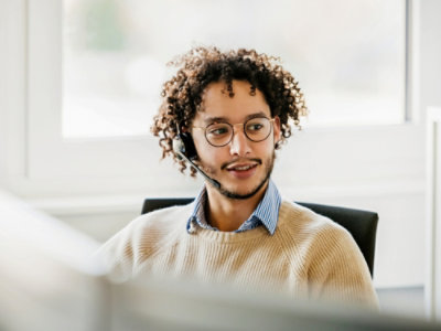 Person wearing a light-colored sweater and headphones, seated at a desk in a bright office environment with a window in the background.