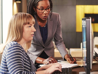 Two people working together at a desk in an office setting. One person is seated, looking at a computer monitor, while the other stands beside them, pointing at a document on the desk.