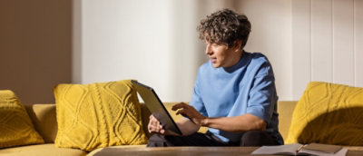 A person sitting on a couch using a laptop with a yellow pillow nearby.