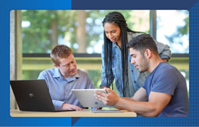 Three people collaborate using a laptop and tablet at a table, reviewing content together in a bright workspace.