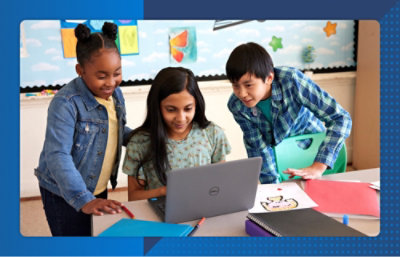 Three students gather around a laptop in a classroom, working together on an activity with notebooks and drawings nearby.