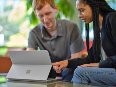 Two people use a laptop together at a table, collaborating in a bright indoor workspace.