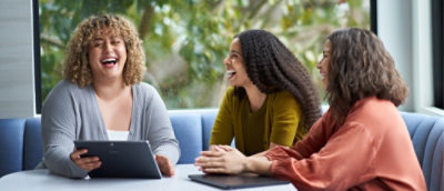 Three people sit together using a tablet, laughing and collaborating in a casual meeting space.