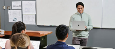 Instructor presents from a laptop at the front of a classroom while students follow along on their own devices.