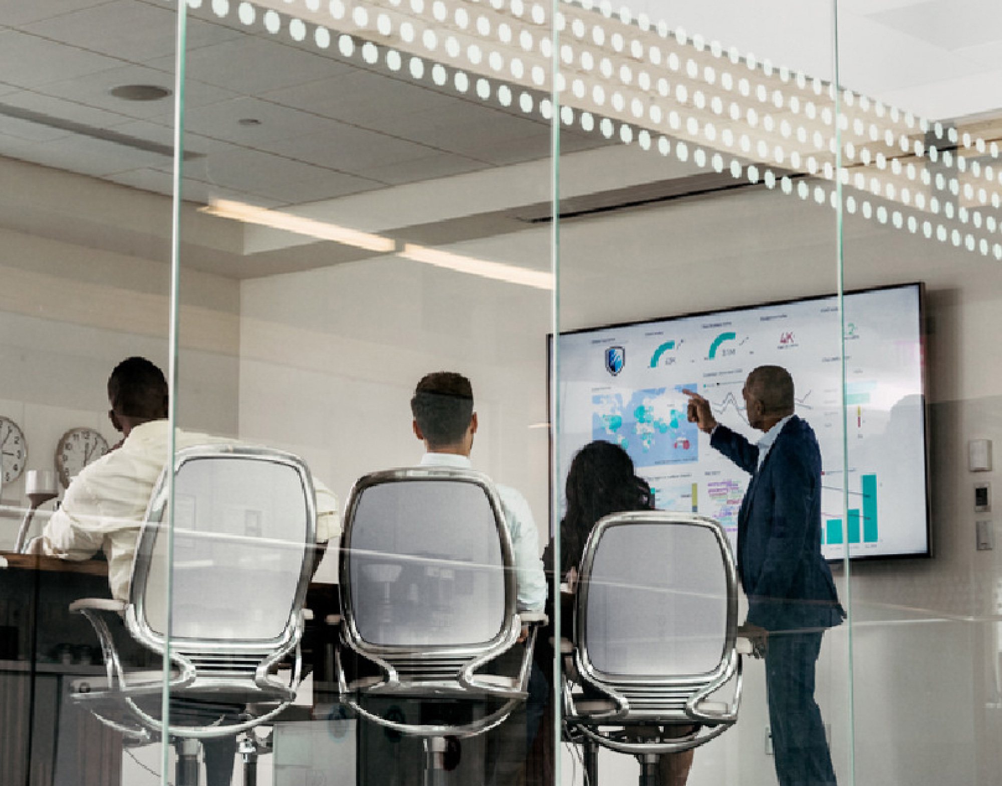 透过玻璃墙看到会议室中有四名专业人员，其中一位指向大屏幕上显示的数字数据。