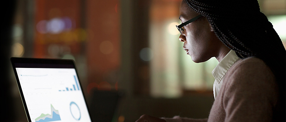 A woman wearing glasses focuses intently on analyzing graphs on her laptop screen in a dimly lit room.