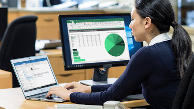 Човек, който пише на лаптоп, свързан с настолен монитор, показващ електронна таблица в Excel