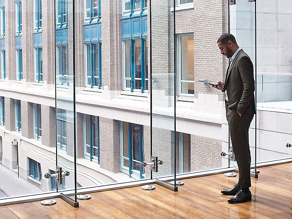 A businessman in a suit checks his smartphone by a glass barrier overlooking a city street with office buildings.