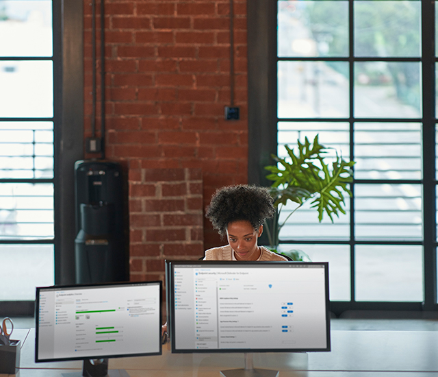 A woman focuses on dual monitors at a desk in an industrial-style office. 