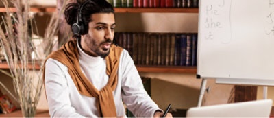 Een persoon met een koptelefoon zit aan een bureau met een notitieblok, pen en een whiteboard met tekst op de achtergrond.