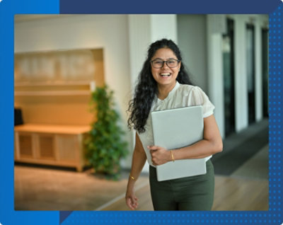 Eine Frau mit langem Haar und Brille lächelt während sie einen zugeklappten Laptop hält und durch einen Bürobereich geht.