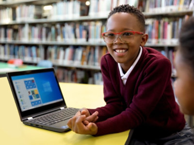 Ein lächelndes Kind sitzt an einem Tisch in einer Bibliothek mit einem aufgeklappten Laptop vor sich. Im Hintergrund sind Bücherregale zu sehen.