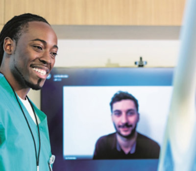Person in medical scrubs standing in front of a computer monitor displaying another individual.