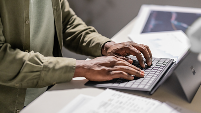 A person types on a laptop at a desk surrounded by scattered documents and charts. The spread of papers suggests a busy, focused work environment..