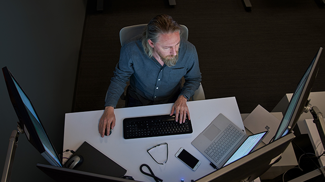 A person with long hair and a beard works at a desk in a dimly lit office. They are using a keyboard and mouse, surrounded by multiple monitors. A laptop, smartphone, glasses, and cables are organized on the desk, creating a tech-focused workspace environment. The setup suggests a professional setting emphasizing technology and multitasking.