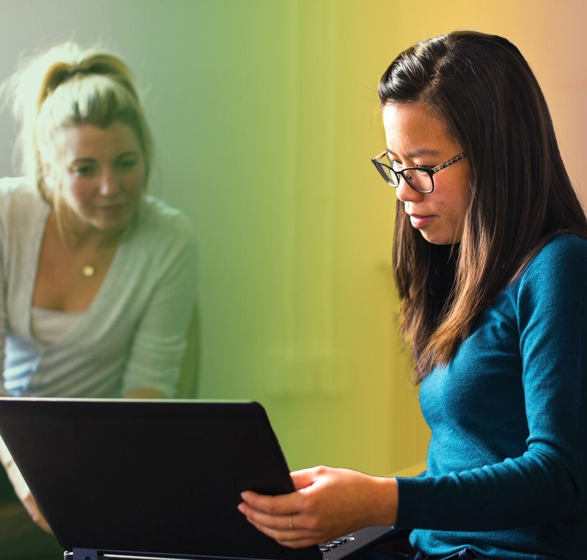 Dwie kobiety, jedna z blond włosami, a druga w okularach, pracują razem na laptopie w kolorowo oświetlonym pomieszczeniu.