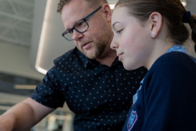 A man with glasses and a young girl focus on something off-frame. The man appears to be explaining or teaching something.