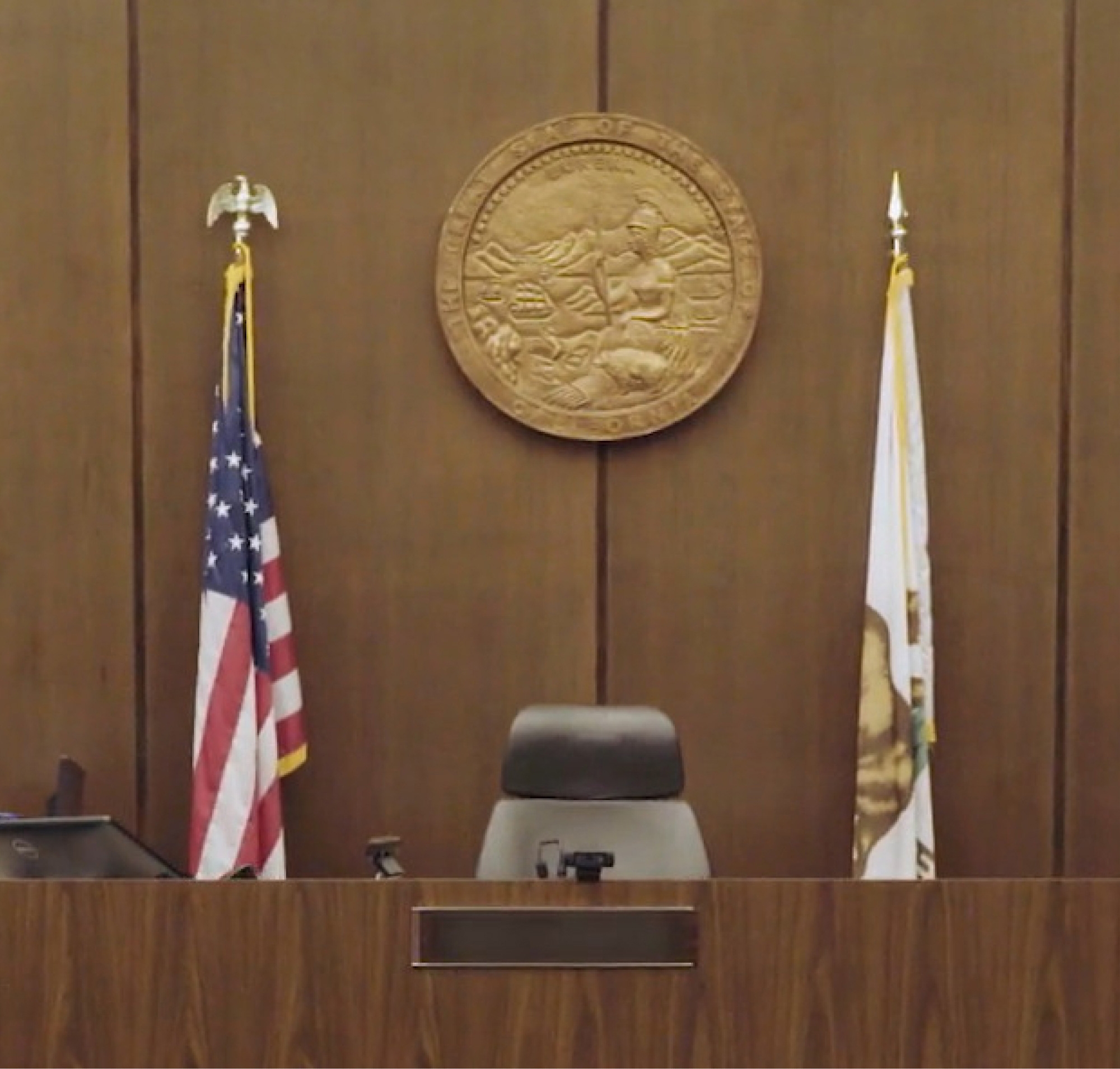 Empty judge's chair in a courtroom with a state seal on the wall, flanked by the u.s. and state flags.
