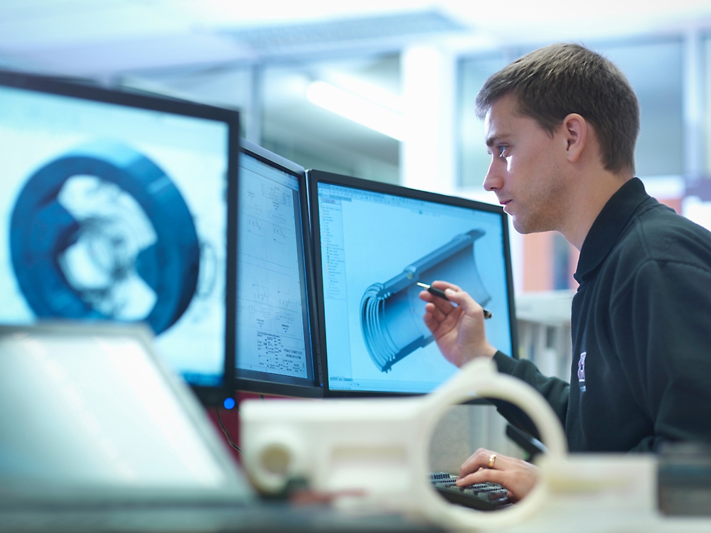 Muž skúma 3D model na obrazovke počítača v technologicky vyspelom pracovisku, drží pero a sústredí sa.