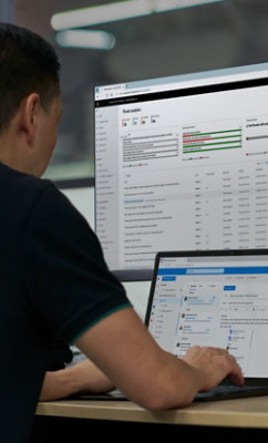 人が机に座ってノート PC で作業しており、その前には大きなコンピューター画面があり、さまざまなシステム データが表示されています