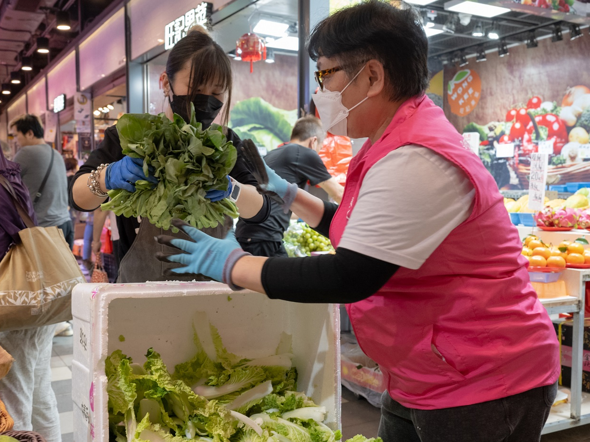 A vendor in a pink shirt and gloves hands fresh vegetables to a woman in a hijab at a busy indoor market. both wear masks.