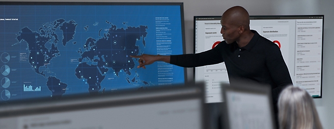 A man points at a digital world map displayed on a screen in a high-tech control room