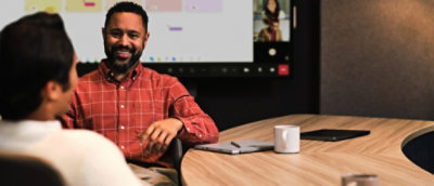 Two professionals discuss at a meeting table, with a large screen showing a video call in the background.