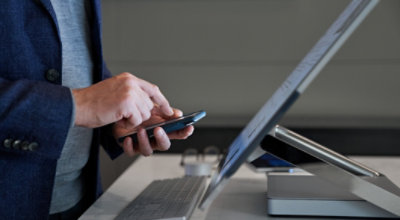 Oseba uporablja telefon z obema rokama in odprt namizni računalnik na mizi