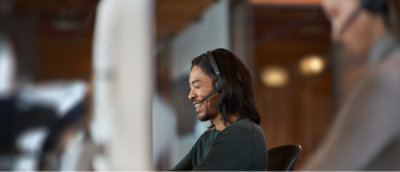A person with long hair, wearing a headset, sits at a desk and smiles while speaking
