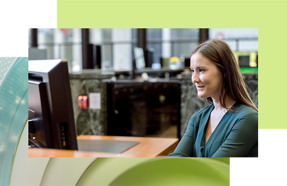 緑色のトップスを着た女性が、オフィス環境でコンピューターを使用しながら、微笑んでいます。
