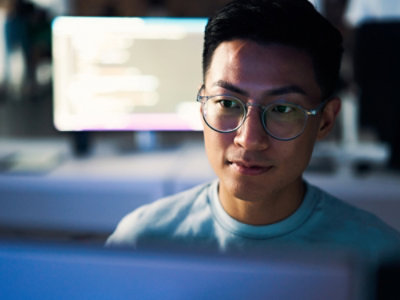 Person wearing glasses working on a screen with blurred code in the background.