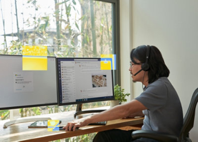 A person sitting at a desk wearing headphones and using a computer with a yellow label on the screen.