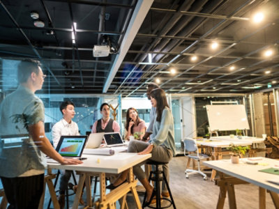 A group of people in a meeting room with a desk, laptop, stool, whiteboard, and a cup on the table.