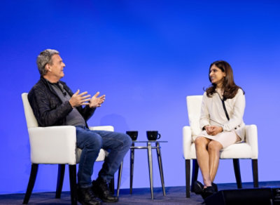 Two people seated on stage in white chairs, talking with a small table and two mugs between them against a blue backdrop.
