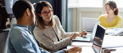 A woman in a white jacket and red glasses sitting at a desk with a laptop.