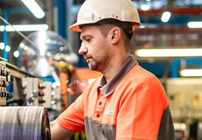 A focused industrial worker wearing a safety helmet and an orange and grey polo shirt inspects machinery in a factory setting
