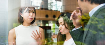 Three people engaged in conversation by a window, with reflections of greenery visible on the glass.