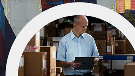 A bald man in a blue shirt using a tablet in a warehouse surrounded by boxes.