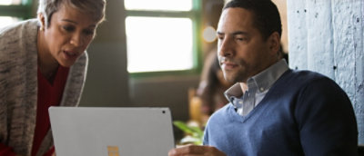  Twee personen kijken samen naar een laptopscherm in een goed verlichte ruimte. De ene zit en draagt een blauwe trui; de andere staat en draagt een jas met een licht patroon.