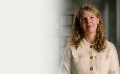 A woman with curly hair wearing cream buttoned shirt, standing indoors against blurred background with soft lighting.