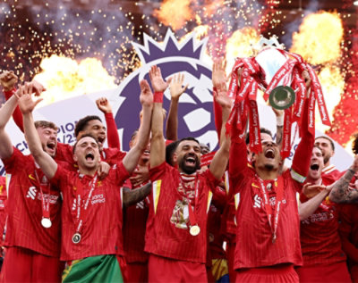 A group of football players celebrating Premier League victory, lifting the trophy high amid roaring flames and confetti, wearing red jerseys with medals.