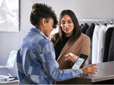 V obchodě s oblečením pomáhá ženě v modré kostkované bundě u pokladny žena v hnědém saku pomocí tabletu.