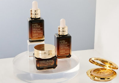 Tři lahvičky Estée Lauder Night Repair, zlatá dóza a pudřenka na průhledném podstavci v elegantním, luxusním prostředí.