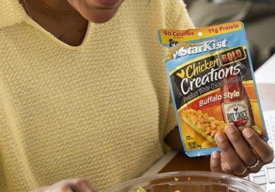 Osoba v žlutém drží nad salátem produkt StarKist Chicken Creations Buffalo Style; na obalu je uvedeno 90 kalorií a 11 g bílkovin.