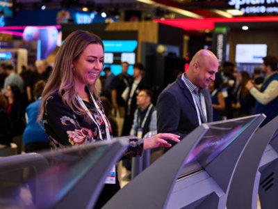 A man and a woman looking at screen and a group of people talking in the background.