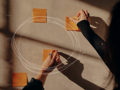 Person arranging orange sticky notes on a wall during a brainstorming session, with handwritten notes, a white circular diagram, and warm sunlight casting shadows.
