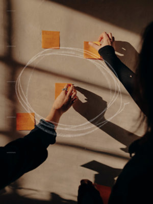 Person arranging orange sticky notes on a wall during a brainstorming session, with handwritten notes, a white circular diagram, and warm sunlight casting shadows.