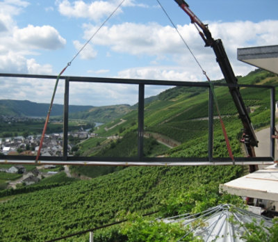 A crane lifting a metal frame over a green field with trees and hills in the background.
